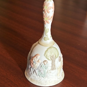 May include: A white ceramic bell with a long handle decorated with peach-coloured flowers and green leaves. The bell features a relief scene of figures in pastel colours, including a tree and people. The bell is a decorative item.