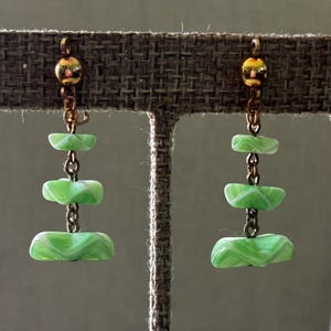 Peut inclure: Une paire de boucles d'oreilles pendantes avec un dessus et une chaîne dorés, comprenant trois perles rectangulaires empilées, vert clair, avec un motif tourbillonnant blanc. Les boucles d'oreilles sont présentées sur un fond de tons neutres.