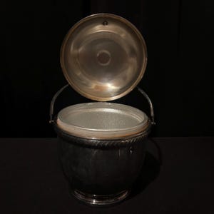 K&ouml;nnte beinhalten: Ein silberner Eisk&uuml;bel mit offenem Deckel, der einen transparenten Glaseinsatz zeigt. Der Eimer hat einen Griff und ein dekoratives, seilartiges Design am Rand. Der Hintergrund ist schwarz.