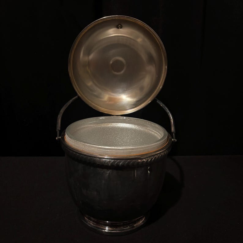 K&ouml;nnte beinhalten: Ein silberner Eisk&uuml;bel mit offenem Deckel, der einen transparenten Glaseinsatz zeigt. Der Eimer hat einen Griff und ein dekoratives, seilartiges Design am Rand. Der Hintergrund ist schwarz.