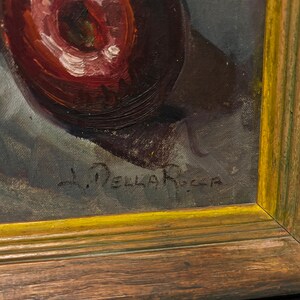 May include: Oil painting of a red fruit, possibly an apple, with white highlights, set against a dark background. The artwork is signed "L. Della Rocca" and framed in a gold-trimmed wooden frame. The painting has a vintage aesthetic.