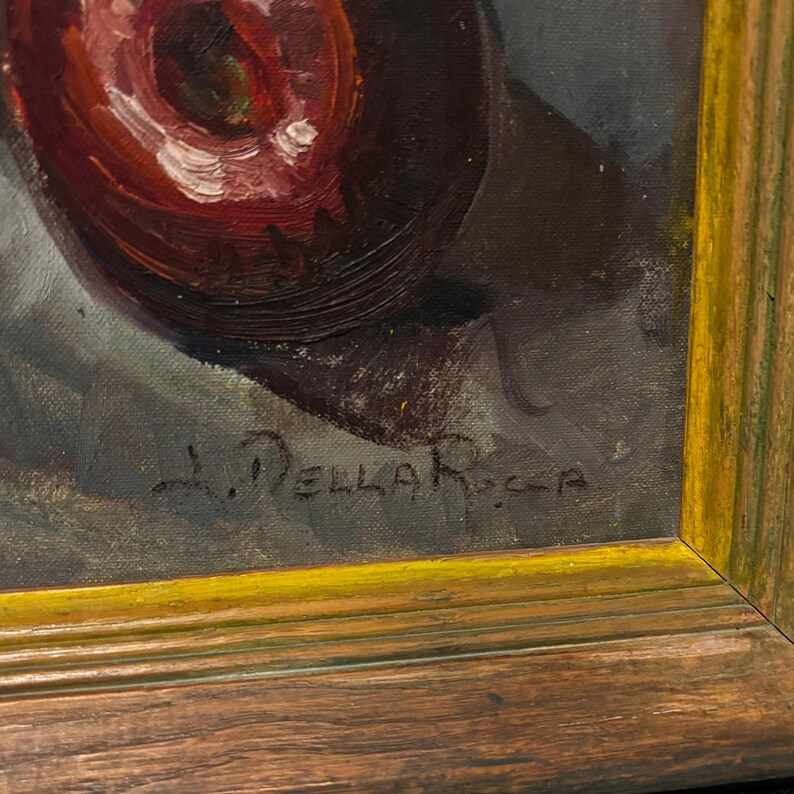 May include: Oil painting of a red fruit, possibly an apple, with white highlights, set against a dark background. The artwork is signed "L. Della Rocca" and framed in a gold-trimmed wooden frame. The painting has a vintage aesthetic.