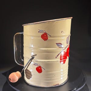 May include: Cream-colored vintage sifter with a handle and a wooden knob. The sifter features red apple illustrations with green leaves. The metal sifter has a cylindrical shape and a handle on the side. The sifter is likely used for baking.