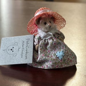 May include: A small teddy bear doll wearing a pink floral dress, a white lace collar, and a red straw hat. A light blue heart-shaped patch is sewn on the dress. A gray tag with the text "Deb Canham Artist Designs Inc." is next to the doll.
