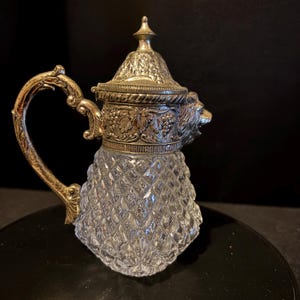 Könnte beinhalten: Eine antike Glaskaraffe mit einem silberfarbenen Metallgriff, Deckel und Akzenten. Der Körper der Karaffe ist aus klarem, rautenförmig geschliffenem Glas. Der Deckel ist verziert und der Griff hat ein dekoratives Design. Auf der Seite der Karaffe ist ein Löwenkopf abgebildet.