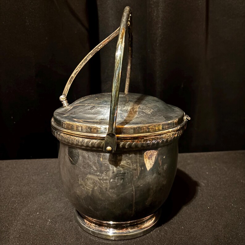 K&ouml;nnte beinhalten: Ein silberfarbener Beh&auml;lter mit Deckel und Henkel. Der Beh&auml;lter hat einen runden K&ouml;rper mit einer dekorativen, seilartigen Verzierung um Deckel und Boden. Der Henkel w&ouml;lbt sich &uuml;ber die Oberseite, und der Deckel ist leicht gew&ouml;lbt.