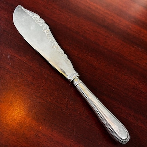 Puede incluir: Cuchillo de mantequilla plateado con un diseño floral decorativo en la hoja. El mango tiene un patrón texturizado y punteado. El cuchillo está sobre una superficie de madera oscura, mostrando su elegante diseño.