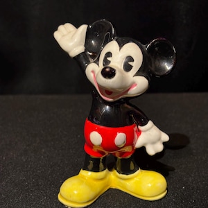 May include: A ceramic figurine of Mickey Mouse, a classic Disney character. Mickey is depicted with a black body, red shorts with white buttons, yellow shoes, and white gloves. He is waving with a smile.