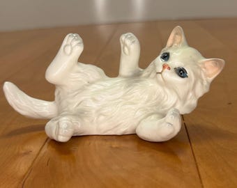 Puede incluir: Una figura de gato de porcelana blanca tumbada boca arriba con las patas levantadas en el aire. El gato tiene pelaje largo esculpido, ojos azul brillante y orejas y nariz con tonos rosados, capturando una pose juguetona con acabado brillante.
