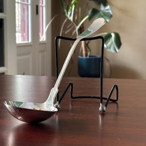 May include: A silver ladle with a rounded bowl rests on a black metal stand. The ladle is positioned on a dark wooden table. A houseplant is visible in the background.