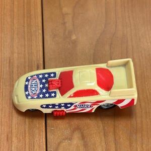 Op de afbeelding: Een speelgoed dragracer in cr&egrave;me, rood en blauw. De auto heeft het NHRA-logo en een patriottisch sterren- en strepenontwerp. De speelgoedauto heeft kleine wielen en een achterspoiler.