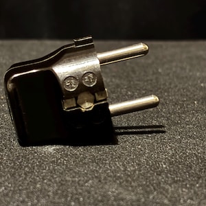 May include: A close-up of a black electrical plug with two silver pins. The plug has a rectangular shape with rounded edges and a small, metallic plate with markings. The background is a dark, textured surface.