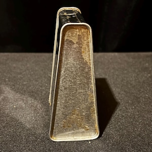 May include: A vintage, silver-colored metal napkin holder. The holder has a rectangular shape with a slightly tapered design. The metal shows signs of age with some discoloration and rust.