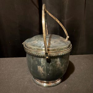 K&ouml;nnte beinhalten: Ein silberfarbener Eisk&uuml;bel mit Scharnierdeckel und Schwinggriff. Der Eimer hat einen runden Boden und eine dekorative, seilartige Verzierung am oberen Rand. Der Griff ist mit kleinen Scharnieren am Deckel befestigt.