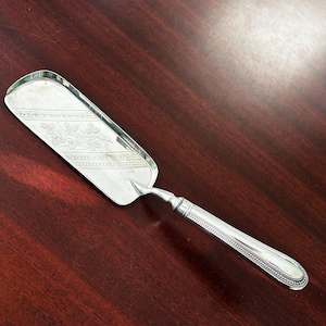 May include: A silver cake server with a rectangular blade and a decorative floral pattern. The handle is detailed with a beaded design. The server is made of a shiny, reflective metal.