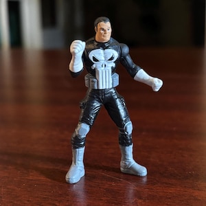 May include: A small action figure of a character in a black and white costume. The figure has a skull emblem on the chest and is posed with one fist clenched. The figure is standing on a wooden surface.