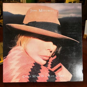 以下が含まれることがあります： ジョニ・ミッチェルのアルバム「Chalk Mark in a Rain Storm」のアルバムカバー。タン色の帽子に黒いバンド、ピンクと白の模様のショールを身につけた女性が写っています。アルバムタイトルとアーティスト名は白で印刷されています。