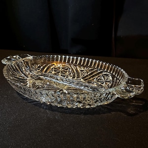 May include: Clear glass serving dish, oval with a divided interior. It has a starburst pattern and handles. The rim has a textured, ridged design. Likely used for snacks or appetizers. The dish measures approximately 25 cm in length.