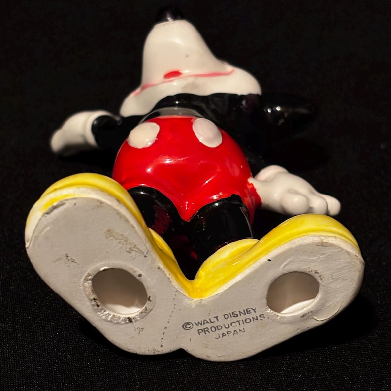 May include: A vintage Mickey Mouse figurine, viewed from below. The figure features a white face, black ears, and a red and white outfit. The yellow shoes are prominent, and the base is marked "Walt Disney Productions Japan."