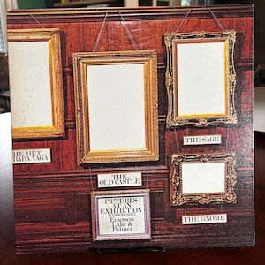 Op de afbeelding: Albumhoes met een donkerhouten achtergrond en verschillende sierlijke gouden fotolijsten. De albumtitel "Pictures at an Exhibition" is zichtbaar, samen met de artiesten Emerson, Lake & Palmer. De frames zijn gelabeld met songtitels.