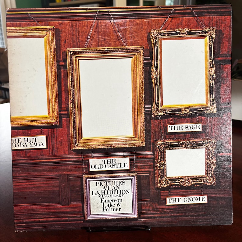 Op de afbeelding: Albumhoes met een donkerhouten achtergrond en verschillende sierlijke gouden fotolijsten. De albumtitel "Pictures at an Exhibition" is zichtbaar, samen met de artiesten Emerson, Lake & Palmer. De frames zijn gelabeld met songtitels.