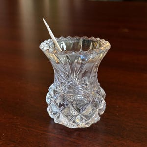 May include: A clear, cut-glass toothpick holder with a decorative, diamond-patterned base and a flared, scalloped rim. A single wooden toothpick is inserted in the holder. The holder is on a dark brown surface.