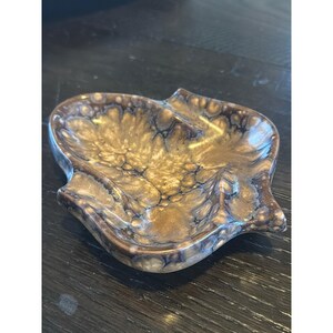 Pode incluir: Um pequeno prato de cerâmica em forma de folha com um esmalte brilhante num padrão marmoreado em tons de dourado metálico, bronze e castanho escuro. O prato tem uma superfície lisa e curva com bordas elevadas e uma textura veada distinta.