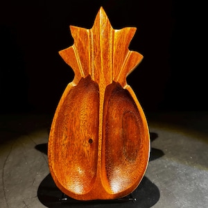 May include: A handcrafted wooden serving dish in the shape of a pineapple. The dish is made of a rich, reddish-brown wood and features two deep, oval-shaped compartments. The top of the dish is carved to resemble the pineapple's crown.