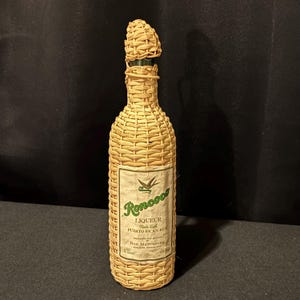 Könnte beinhalten: Eine Vintage-Flasche Roncoo-Likör, hergestellt mit puerto-ricanischem Rum, ist in geflochtenem Stroh eingeschlossen. Die Flasche hat einen passenden strohbedeckten Verschluss. Das Etikett zeigt grünen Text und eine Vogelgrafik.