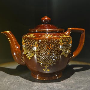 May include: A brown ceramic teapot with a lid and handle. The teapot is decorated with a floral pattern in yellow, orange, and white. The spout and handle are curved. The teapot sits on a dark surface.