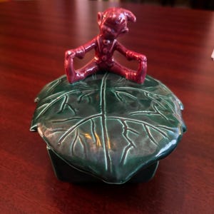 Puede incluir: Caja de baratijas de cerámica con tapa en forma de hoja verde oscuro. Una pequeña figura de elfo de color burdeos se encuentra encima de la tapa. La caja tiene una base cuadrada y probablemente sea vintage. El diseño general es caprichoso y decorativo.