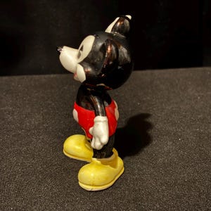 May include: A vintage Mickey Mouse figurine. The figure has a black head and body, white gloves, red shorts with white buttons, and yellow shoes. The figurine is a classic representation of the iconic cartoon character.
