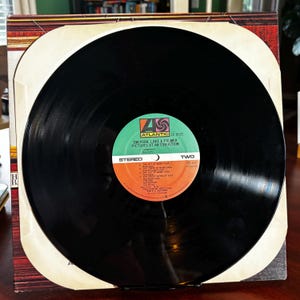 Op de afbeelding: Een zwarte vinylplaat met een groen en oranje label. Het label leest "EMERSON, LAKE & PALMER PICTURES AT AN EXHIBITION" en "STEREO". De plaat zit in de originele albumhoes.