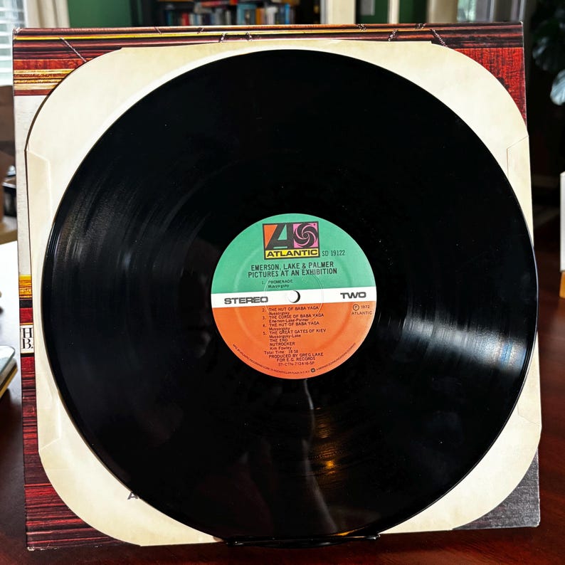 Op de afbeelding: Een zwarte vinylplaat met een groen en oranje label. Het label leest "EMERSON, LAKE & PALMER PICTURES AT AN EXHIBITION" en "STEREO". De plaat zit in de originele albumhoes.