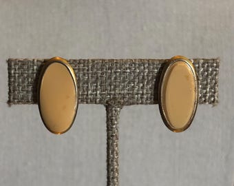May include: A pair of clip-on earrings. Each earring features an oval, cream-colored center with a gold-colored border. The earrings are displayed on a textured, gray fabric.