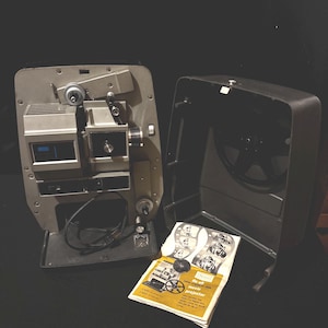 Puede incluir: Proyector de películas vintage con cuerpo de metal plateado y estuche negro. El proyector está abierto, revelando sus componentes internos. Un folleto con ilustraciones y texto está junto al proyector. El folleto dice "Du-all 8mm movie projector".