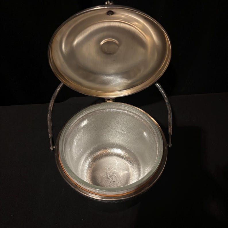 K&ouml;nnte beinhalten: Ein versilberter Eisk&uuml;bel mit Glasinnenraum, offenem Deckel und Griff. Der Eimer ist rund mit einem dekorativen Rand und einem Klappdeckel. Der Griff ist gebogen und an den Seiten des Eimers befestigt. Das Innenglas ist mattiert.