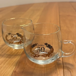 Puede incluir: Dos piezas de servicio de vidrio transparente con ribete dorado. Una es una jarrita con asa y boquilla, la otra es un vaso redondo. Ambas tienen un diseño dorado "Golden 50 Anniversary".