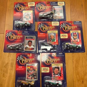 May include: A collection of die-cast model race cars in their original packaging. Each package features a miniature race car, a trading card of John Force, and the "Winner's Circle" logo. The cars are various colors, including black, green, and gold.