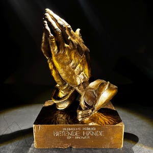 May include: A gold-colored sculpture of praying hands, resting on a rectangular base. The base features the text "ALBRECHT DÜRER'S BETENDE HÄNDE BY BROWER" in a gold tone. The sculpture is set against a dark background.