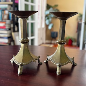 May include: Two antique brass candle holders. Each has a wide, shallow bowl at the top, a geometric column, and a decorative base with three feet. The candle holders are a warm, aged brass color.