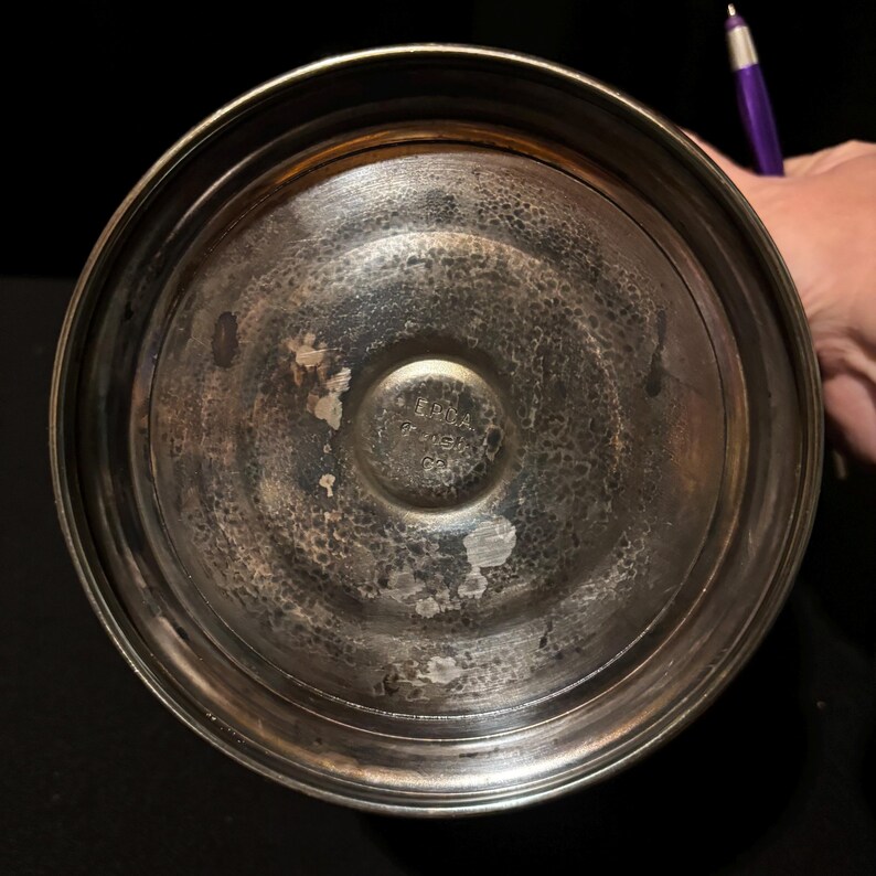 K&ouml;nnte beinhalten: Nahaufnahme des Bodens eines silberfarbenen Metallbeh&auml;lters. Das Metall hat ein strukturiertes, geh&auml;mmertes Aussehen mit Abnutzung und Verf&auml;rbung. Der Boden hat einen schwer lesbaren Stempeltext. Der Beh&auml;lter steht vor einem dunklen Hintergrund.