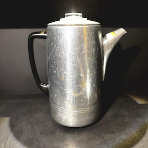 Può includere: Una caffettiera vintage color argento con un manico nero e un coperchio trasparente e rotondo. La caffettiera ha un beccuccio ed è realizzata in metallo. La superficie mostra segni di usura, suggerendo che si tratta di un oggetto più vecchio. La caffettiera è probabilmente per preparare il caffè.