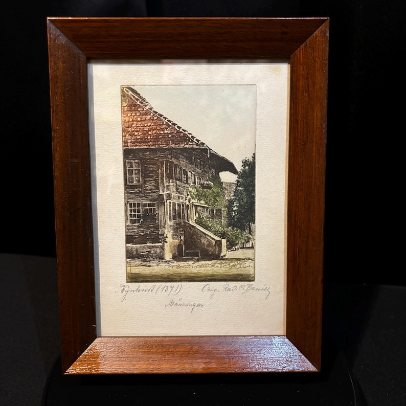 May include: A framed print depicting a historic building with a red tiled roof and wooden facade. The print is set within a dark wood frame. Text is visible at the bottom of the image.