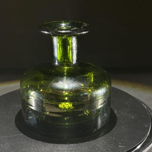 May include: A green glass bottle stopper with a rounded base and a short, cylindrical neck. The stopper has a flat, circular top. The glass has a slight green tint and reflects light, creating highlights and shadows. The object is likely for a wine bottle.