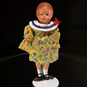 Puede incluir: Muñeca vintage con rostro pintado y pelo corto pelirrojo. La muñeca viste un vestido amarillo con estampado floral, cuello blanco con ribete azul marino y zapatos negros. Una etiqueta dorada en forma de corazón cuelga de una cadena alrededor del cuello.