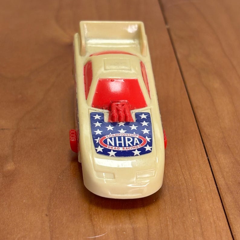 Op de afbeelding: Een vintage speelgoed dragraceauto in cr&egrave;me, rood en blauw. De auto heeft de tekst "NHRA Drag Racing Championship" op de motorkap, met een sterrenpatroon. De wielen en andere details zijn rood.