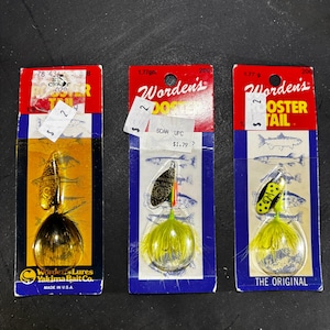 May include: Three fishing lures on their original packaging. The lures are Worden's Rooster Tail brand, with a variety of colors and designs. The packaging is red, blue, and yellow, with the brand name prominently displayed. The lures are designed for catching fish.