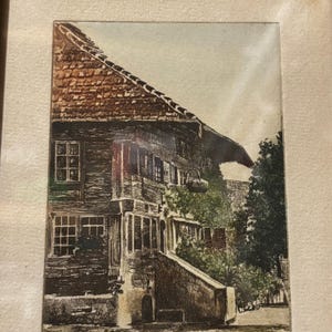 May include: A framed print depicting a traditional building with a tiled roof. The print features a detailed illustration of the building's facade, windows, and a stone staircase. The artwork is in shades of brown, green, and beige, with a vintage aesthetic.
