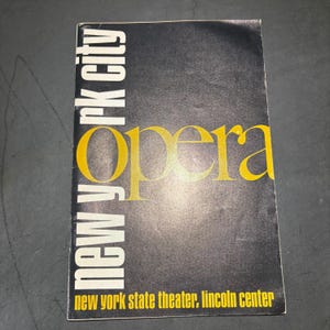 Puede incluir: Un programa de ópera vintage de la ciudad de Nueva York con un fondo negro. La palabra "Opera" está en dorado, y "New York City" está en blanco. El texto "New York State Theater, Lincoln Center" está en la parte inferior.